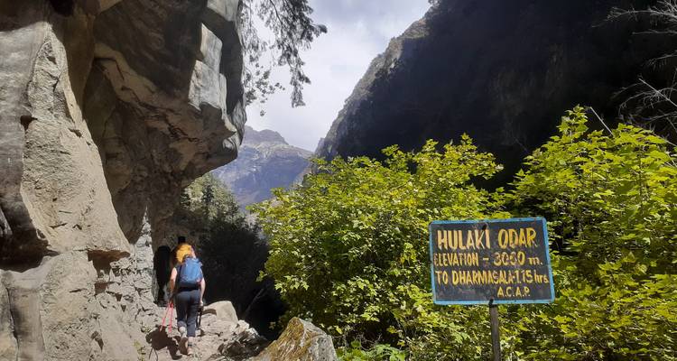 Hiker walking on a trail near a rocky cliff with a sign for Hulaki Odar