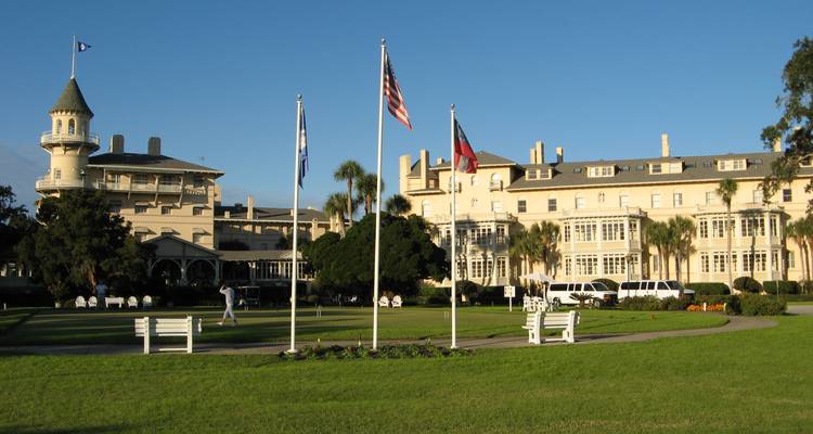 Pelouse et mâts de drapeaux devant le complexe hôtelier historique du Jekyll Island Club sous un ciel bleu dégagé.