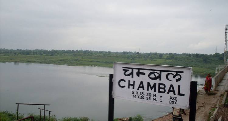 Sign reading 'CHAMBAL' beside a wide river under overcast sky with a lone person walking
