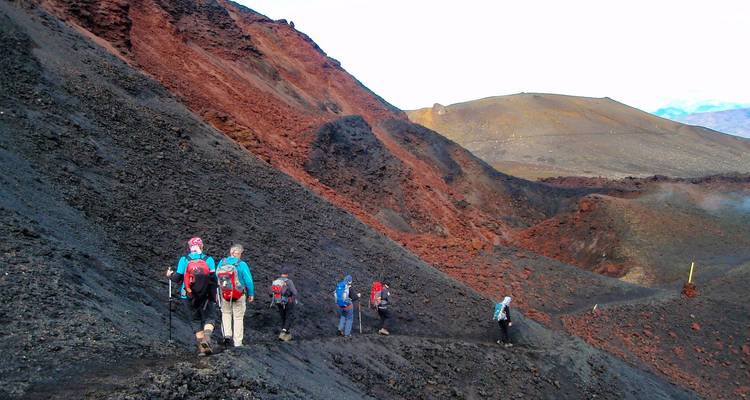 Hikers traverse a steep trail across dark volcanic slag with red rock ridges all around.