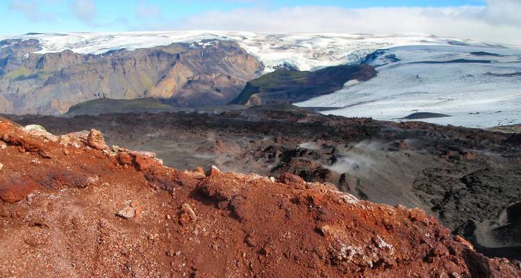 Steam rises from black volcanic ground with red rocks in the foreground and a sweeping glacier beyond.