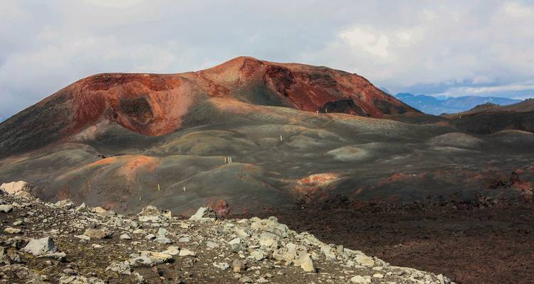 Colorful volcanic cone rises above barren lava fields under a moody sky.