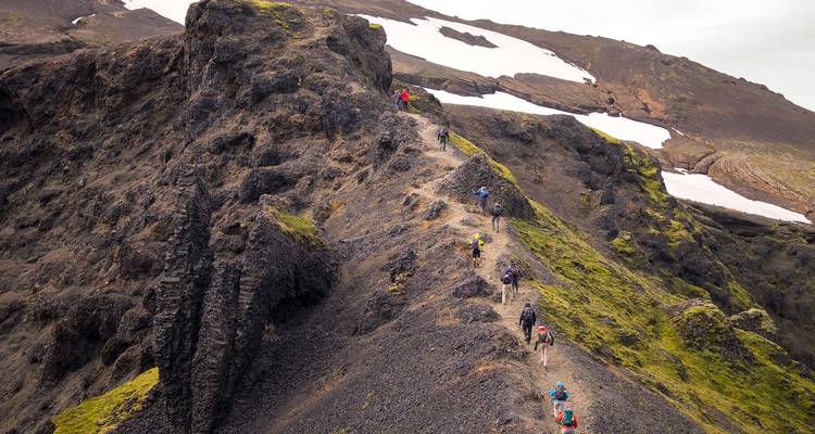 Adventurers hike along a dramatic knife-edge volcanic ridge with snow patches and moss-covered slopes.