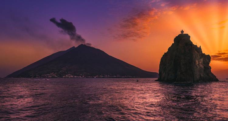 Coucher de soleil dramatique sur une île volcanique avec de la fumée qui s'élève et une petite île avec un phare.