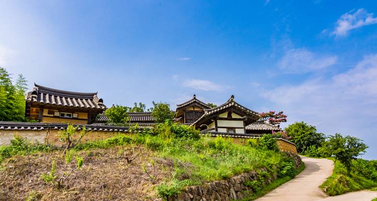 Traditionelle Hanok-Gebäude an einem sanften Berghang mit einem geschwungenen ländlichen Weg unter einem strahlend blauen Himmel.