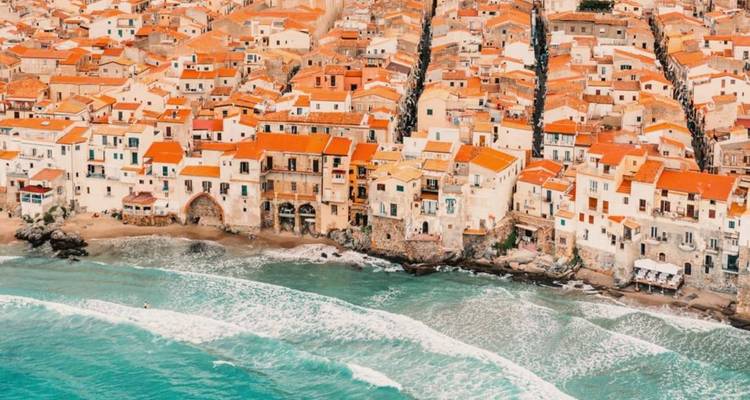 Aerial image of Cefalù’s terracotta rooftops cascading to a narrow beach with waves rolling in.