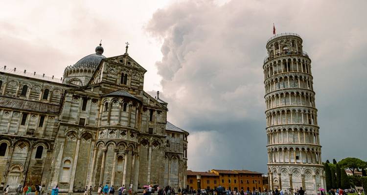 Pisa’s Leaning Tower and cathedral stand against dramatic storm clouds over a busy square.