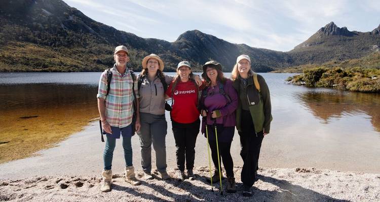 Cinq randonneurs heureux posent ensemble sur une plage de galets avec un lac alpin limpide et des collines escarpées en arrière-plan.