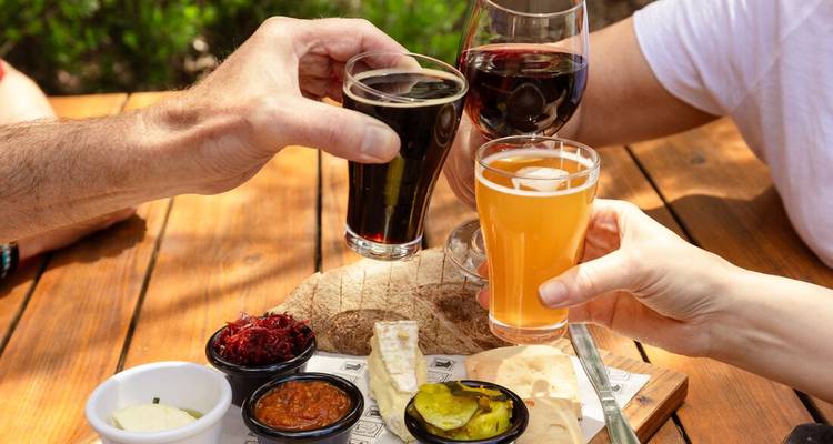 Hands toast with craft beer and wine over a wooden table spread with local cheeses and chutneys.