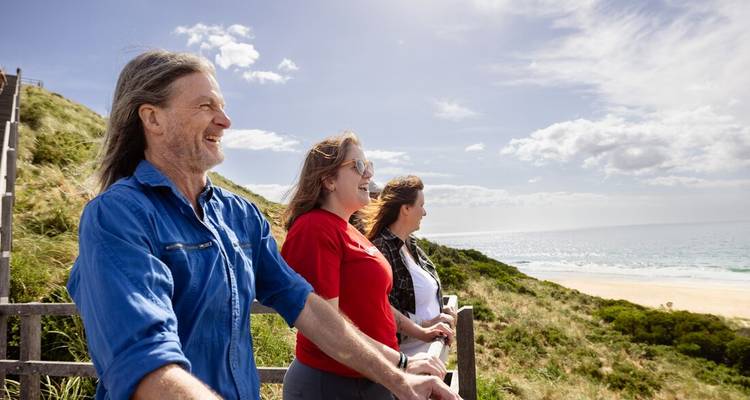 Travellers smile as they enjoy ocean views from a clifftop lookout on a bright, breezy day.