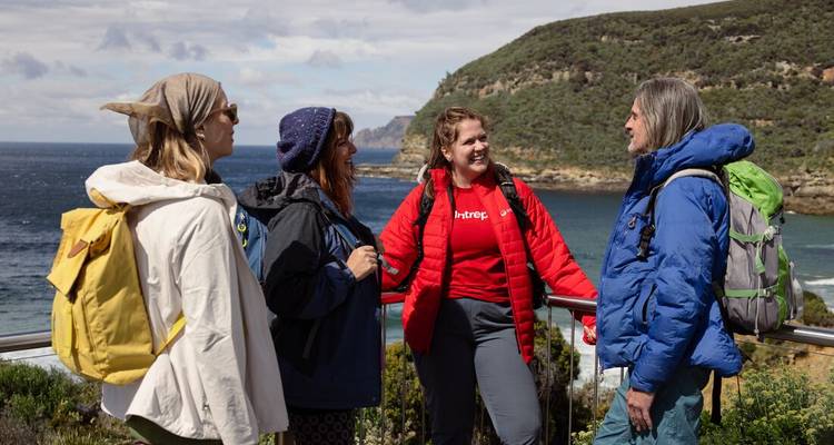 Small group chats at a coastal railing with rugged headlands and blue sea behind them.