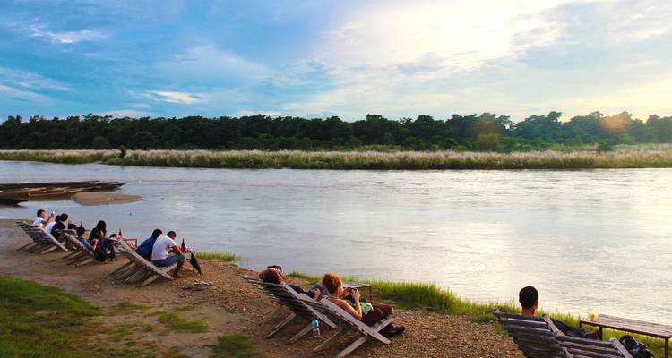 People relaxing on chairs by a river bank during sunset.