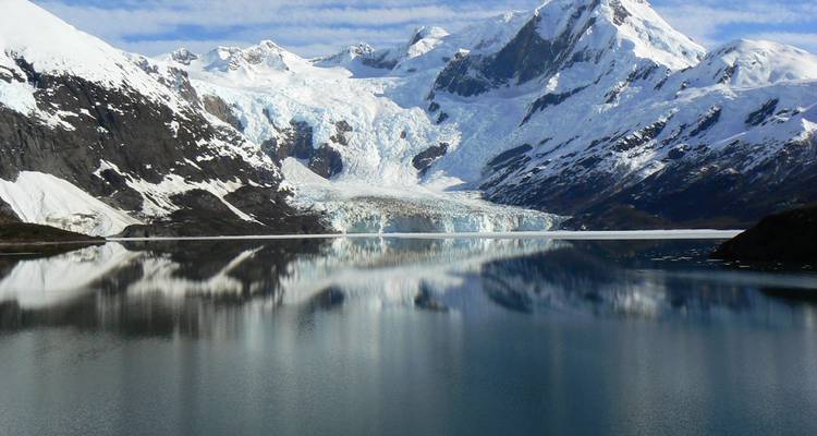 Lac glaciaire lisse comme un miroir reflétant les pics enneigés imposants et les glaciers sous des nuages striés.