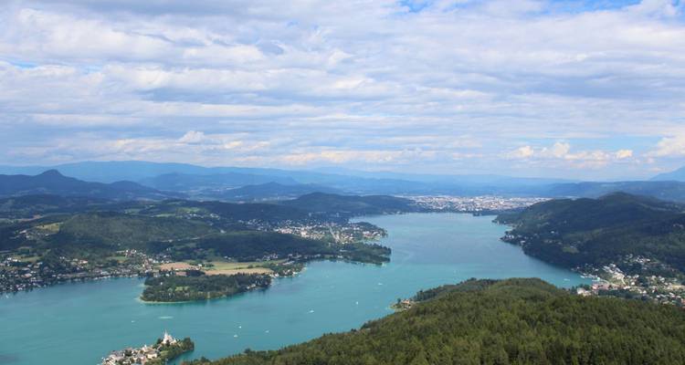 Expansive lake bordered by forested hills and distant mountains under a partly cloudy sky.