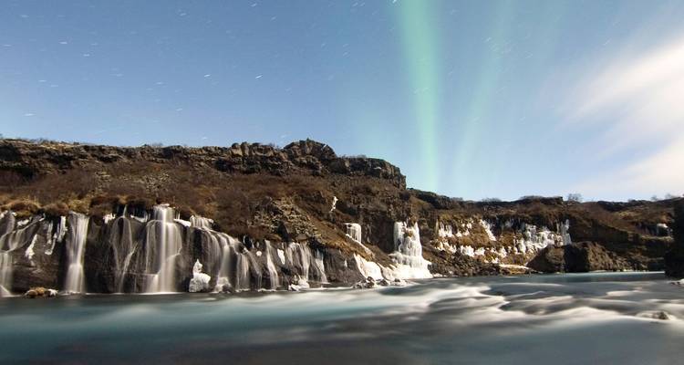 Long-exposure image of Icelandic waterfalls under a sky glowing with aurora borealis.