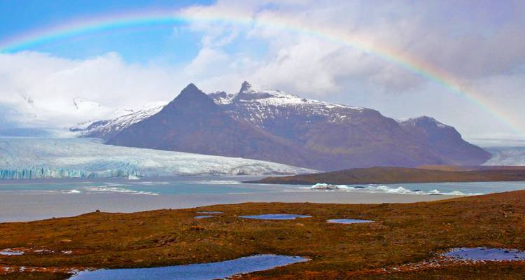 Vivid rainbow arches over dramatic glacier-capped mountains and icy lagoon pools in south Iceland.