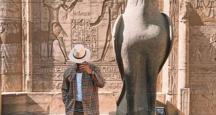 Mann mit Hut und kariertem Hemd posiert vor antiken Schnitzereien und einer Falkenstatue.