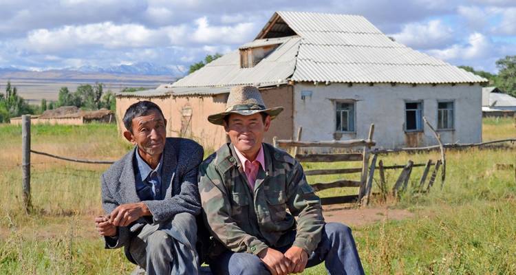 Two people sitting in front of a rustic house with mountainous scenery.