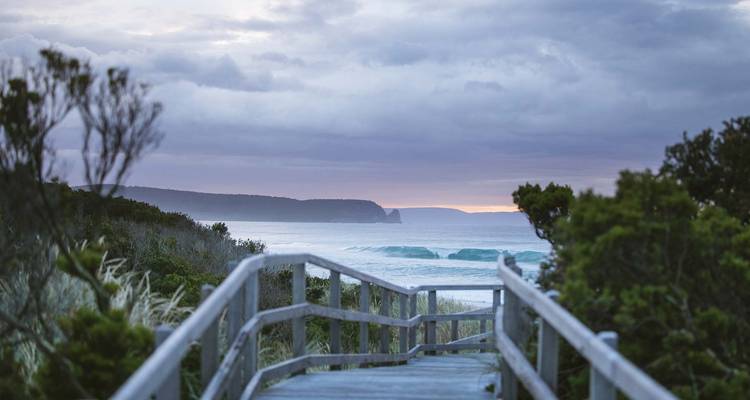 Holzsteg, der zu einem Strand mit Wellen und bewölktem Himmel führt.