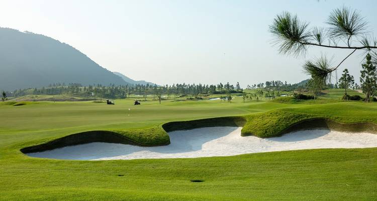 Een groene golfbaan met zandbunker en heuvels in de verte.