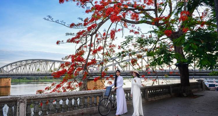 Zwei Frauen in traditioneller Kleidung posieren mit einem Fahrrad an einem Fluss, mit einer Brücke und einem rot blühenden Baum im Hintergrund.