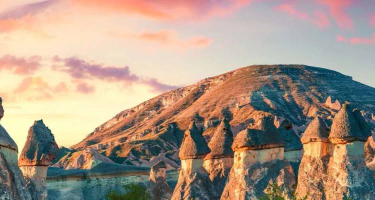 Fairy-chimney rock formations glow under a vivid pink-orange sunset sky with a conical hill rising behind in Cappadocia.