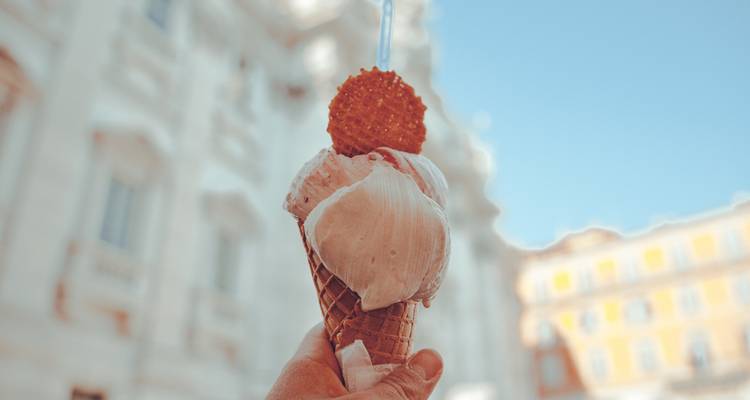 Gros plan d'une main tenant une glace crémeuse dans un cornet gaufré avec des bâtiments historiques flous en arrière-plan.