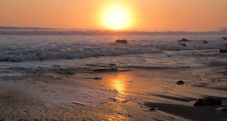 Sonnenuntergang über dem Ozean mit Wellen und Felsen am Ufer.