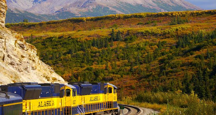 Un train jaune vif de l'Alaska Railroad serpente à travers une vallée de feuillage automnal sous des pics escarpés.