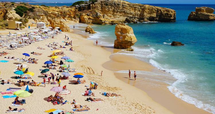 Una playa de arena con personas relajándose bajo sombrillas y rocas al fondo.