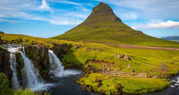 Das ikonische Berg Kirkjufell erhebt sich über einen kleinen Wasserfall an einem hellen Sommertag.