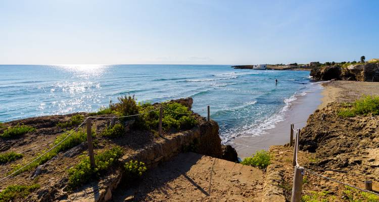 Playa rocosa y tranquila con olas suaves y un nadador solitario bajo el sol brillante.
