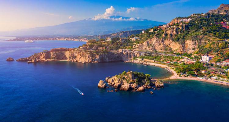 Espectacular panorama aéreo de la costa de Taormina con el Monte Etna elevándose a lo lejos bajo un cielo azul.