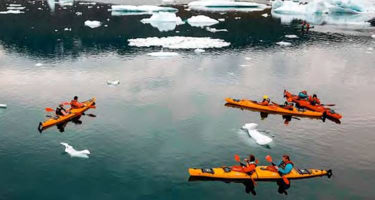 Kayakers maneuvering between icebergs in a cold, scenic setting.