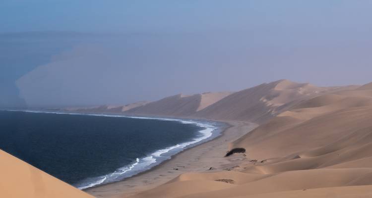 Dunas de desierto que se extienden hasta el Océano Atlántico con una costa curva bajo un cielo azul brumoso