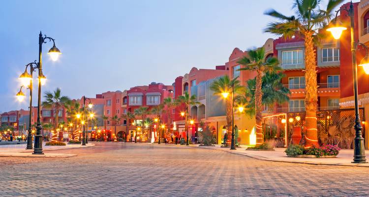 Scène de rue en soirée de la promenade moderne de Hurghada avec une avenue bordée de palmiers illuminés et des bâtiments aux couleurs vives.