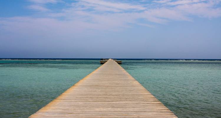 Une longue passerelle en bois pointe vers la mer Rouge turquoise et cristalline sous un ciel légèrement nuageux.