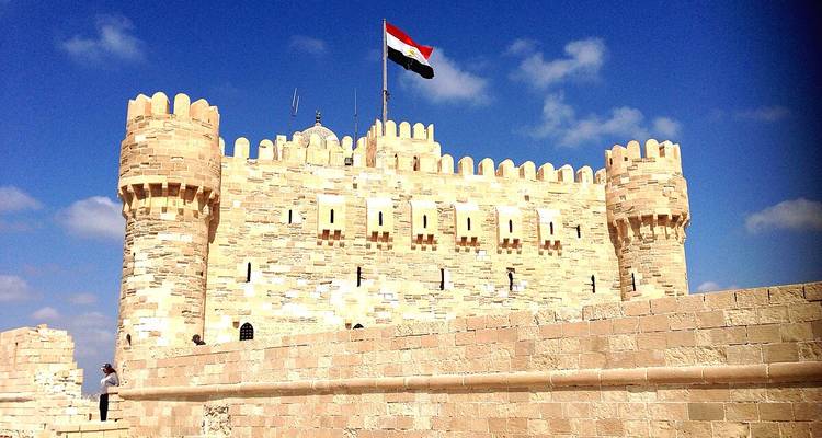 Murs de pierre et tours crénelées de la Citadelle de Qaitbay arborant le drapeau égyptien contre un ciel dégagé.