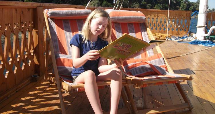 Jeune fille lisant un livre tout en se relaxant sur une chaise longue rayée sur le pont d'un bateau en bois.