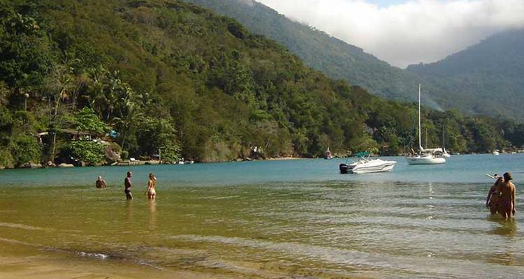 Mensen genieten van een strand met boten en groene heuvels op de achtergrond.