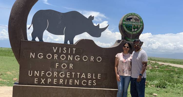 Ein Paar posiert vor dem geschnitzten Nashorn-Willkommensschild in Ngorongoro unter einem strahlend blauen Himmel.