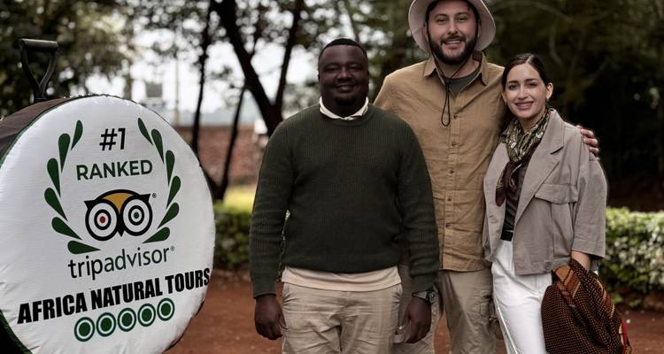 Drei Reisende lächeln neben einer großen TripAdvisor-Auszeichnungstrommel an einem Safari-Treffpunkt.