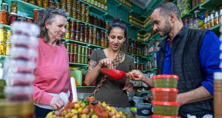 Tourists sample olives and pickles inside a colourful Moroccan souk lined with glass jars.