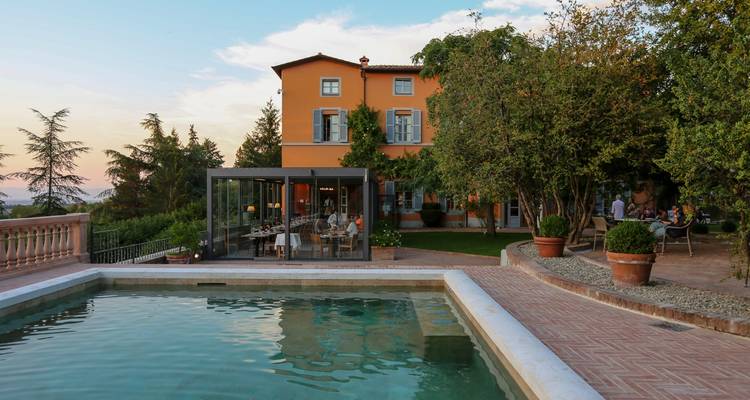 Elegant country villa with pool, glass dining pavilion, and guests enjoying the garden at dusk.
