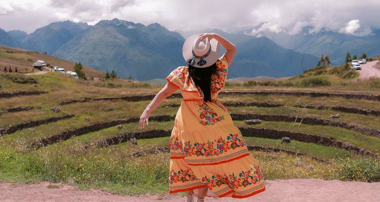 A woman in a flowing orange dress twirls gracefully overlooking the circular agricultural terraces of Moray with misty Andes behind.