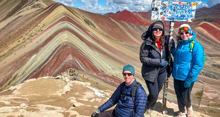 Des voyageurs souriants à côté d'un panneau de direction marquant le sommet de la Montagne Arc-en-ciel avec une crête aux couleurs vives en arrière-plan.