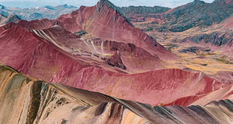Vue aérienne panoramique des montagnes andines aux teintes variées avec des crêtes spectaculaires rouges et beiges sous un ciel dégagé.