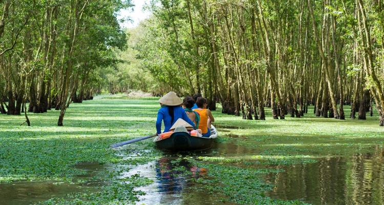 People rowing a boat through a green, tree-lined waterway.