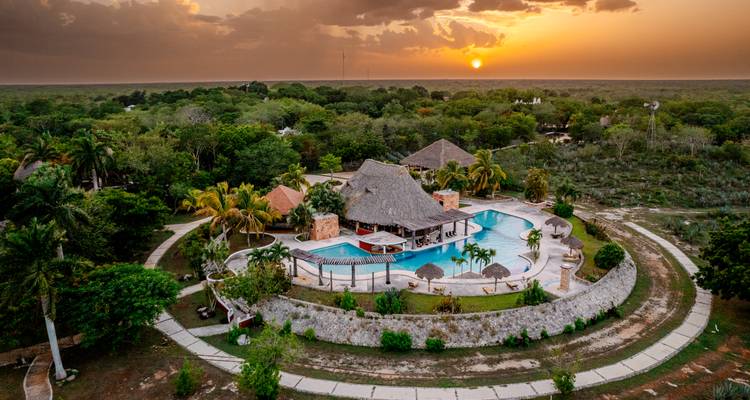 Vista aérea al atardecer de un resort tropical con piscina rodeada de palmeras en medio de la selva.