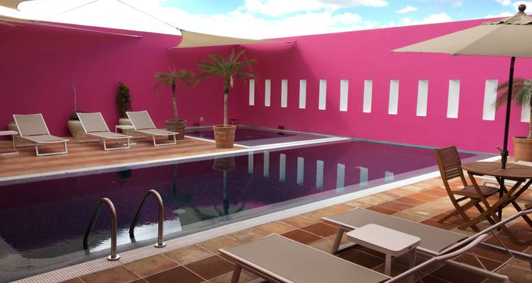 Zone de piscine d'un hôtel de charme avec des murs rose vif, des chaises longues, des parasols et des jardinières avec palmiers.